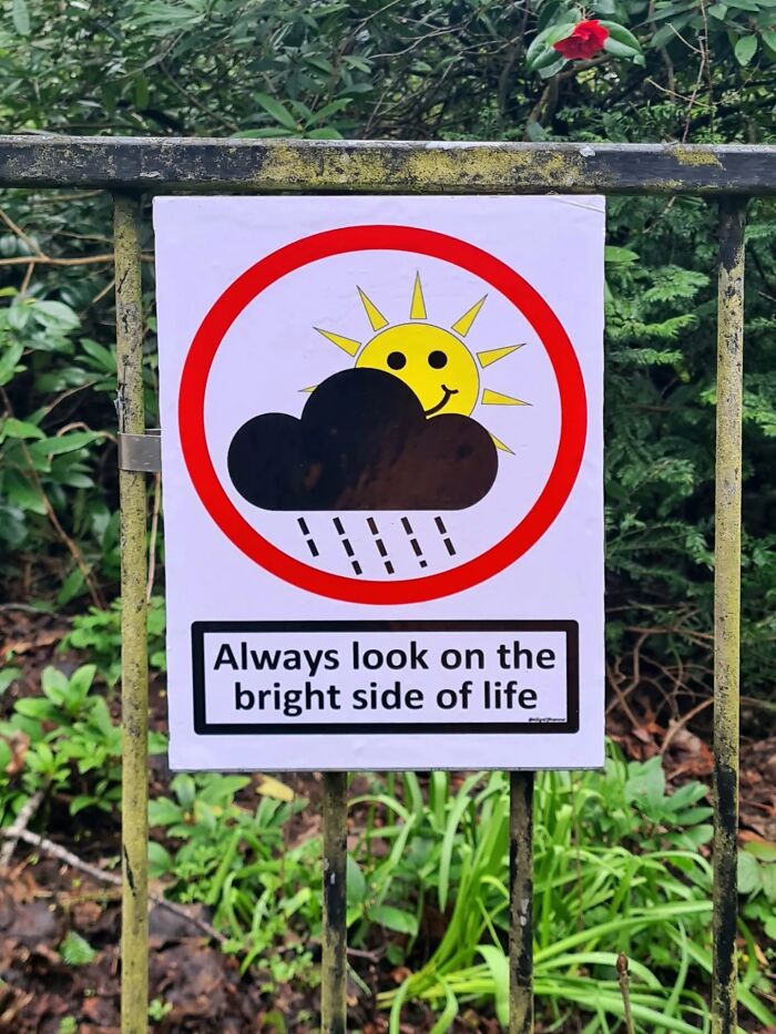 A brilliantly absurd sign showing a sun behind a rain cloud, with text encouraging optimism.
