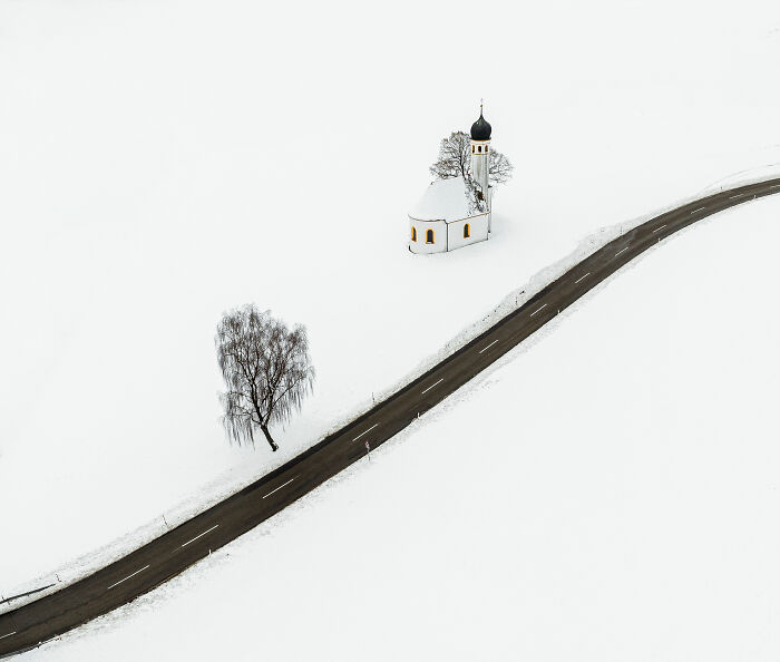 Spectacular aerial photograph of a solitary church and winding road in a snow-covered landscape, awarded at the Aerial Photographer of the Year competition.