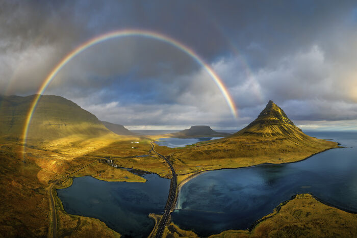 Spectacular aerial photo of a vibrant rainbow over mountains, a road, and water, celebrated at the Aerial Photographer of the Year competition.