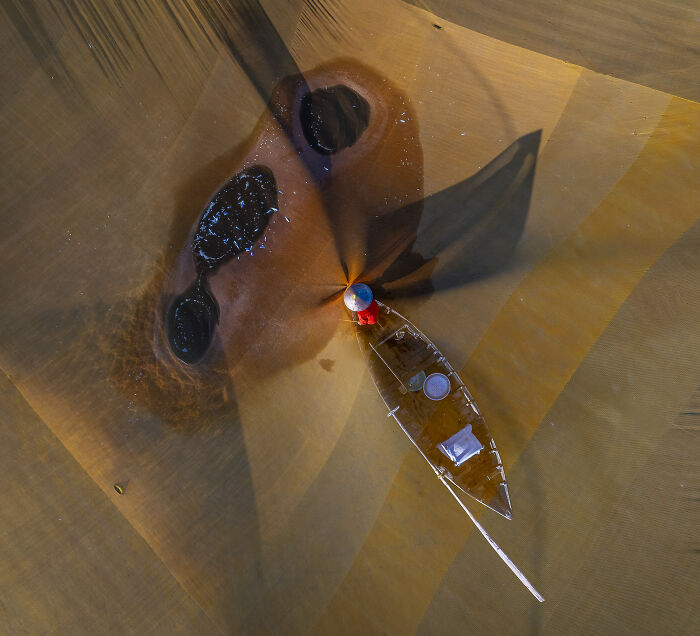 A small boat with a person in a conical hat in shallow, patterned water, an aerial photograph awarded at the 2026 competition.