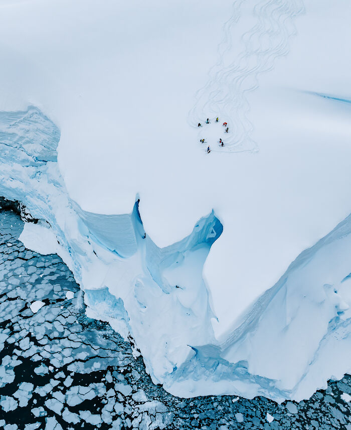 Spectacular aerial photograph of skiers on a snowy iceberg surrounded by icy water, awarded at the Aerial Photographer of the Year competition.