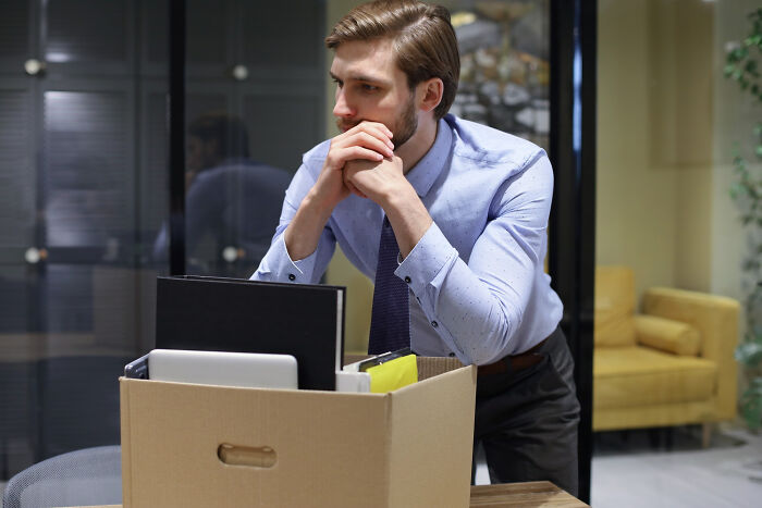 A man with a pensive expression stands next to a box of personal items, signifying dumb reasons people got fired.