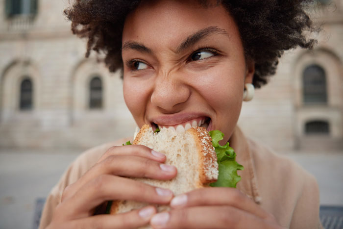 A young, curly-haired student worker intensely biting into a sandwich, representing her struggle with a lazy manager.