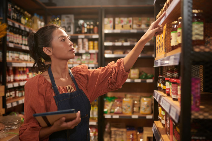 A female student worker in an apron and orange top uses a tablet to check stock in a store, relating to being fired.