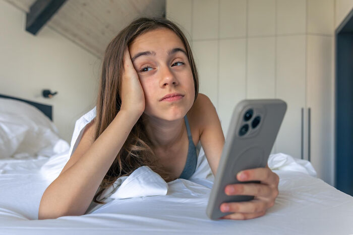 A girl in bed looking annoyed while holding her phone, possibly after a pocket dial incident or avoiding one.