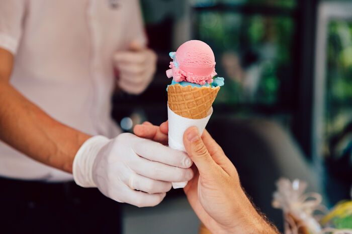 A gloved hand serves a pink and blue ice cream cone to another hand, subtly hinting at dumb reasons for being fired.