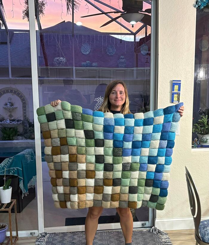 A woman happily holding up a large, colorful checkerboard crochet blanket, an incredible piece by a crochet enthusiast.