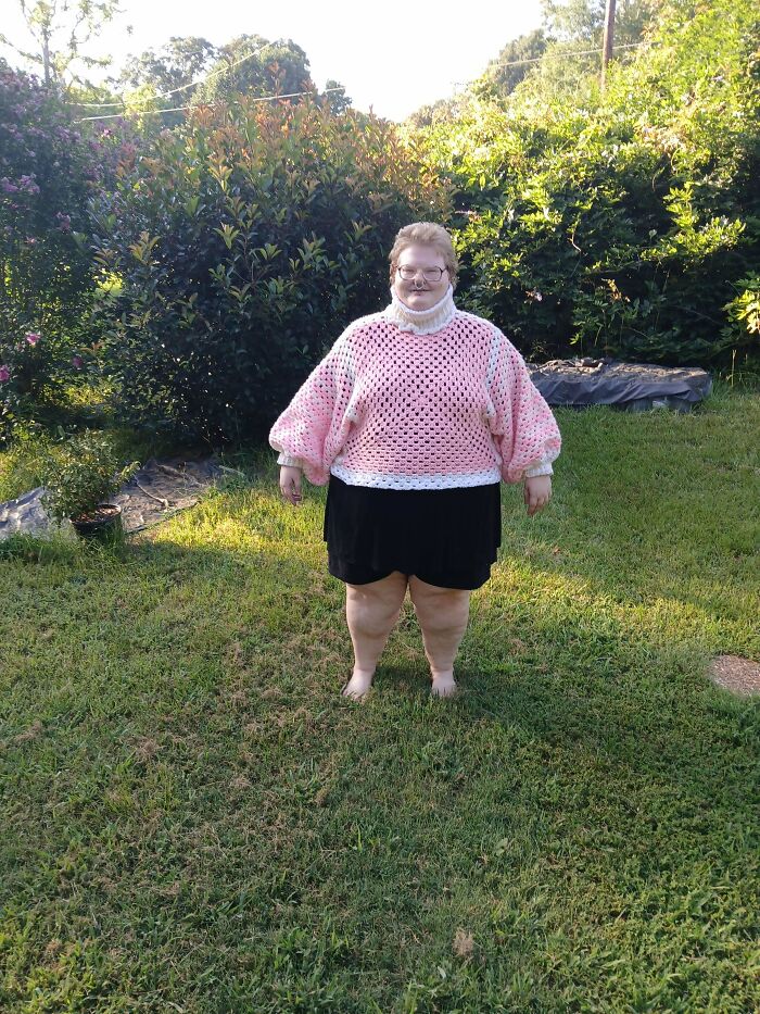 A person stands outdoors, proudly wearing a handmade pink and white crochet sweater, an incredible creation by an enthusiast.