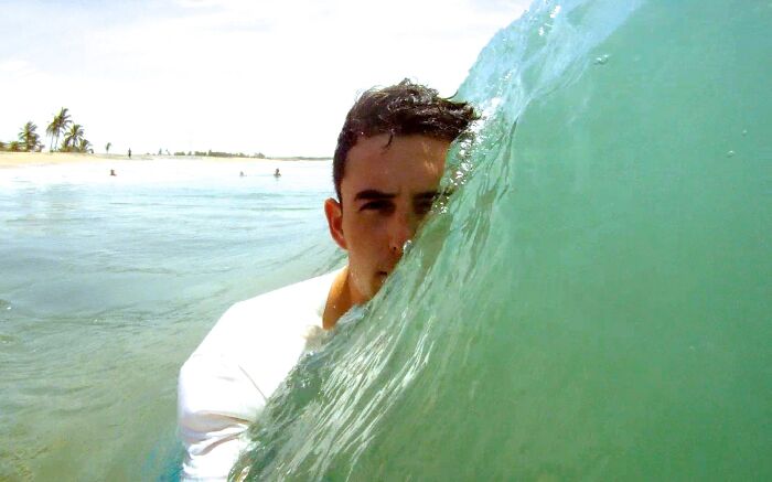 A man's face peeks out from behind a large wave in the ocean, moments before disaster.