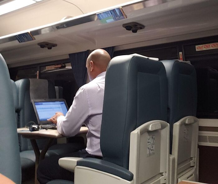 A bald man sitting on a train, working on his laptop. He is one of the entitled men with infuriating stories.
