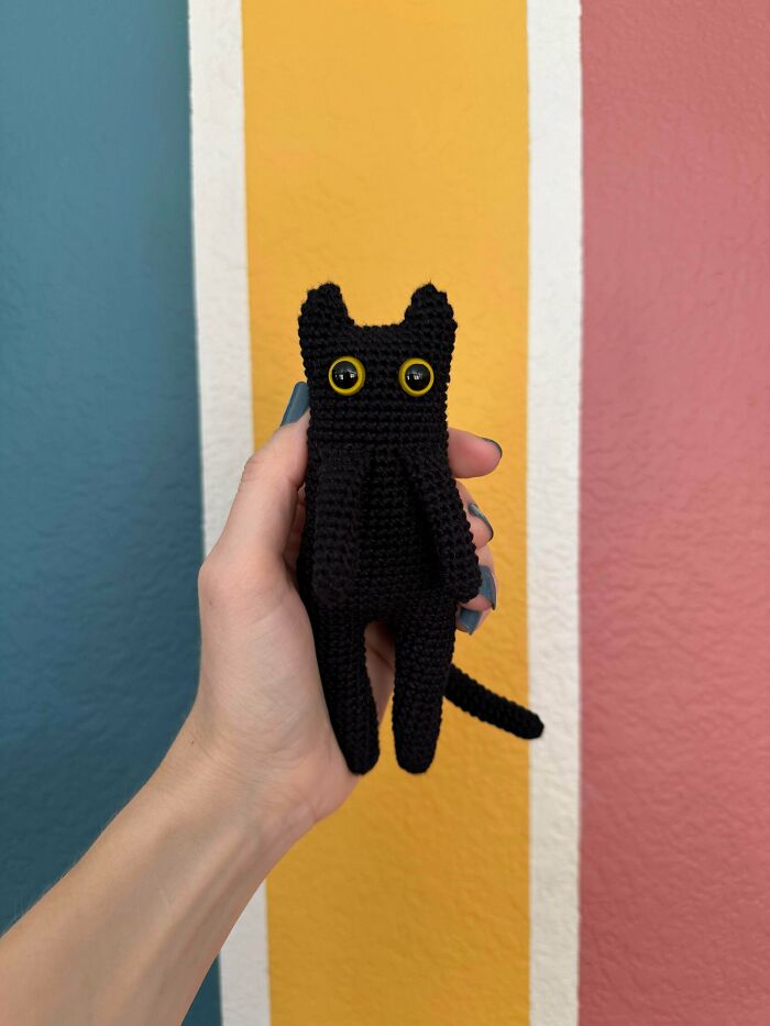 A hand holding a cute, black crochet cat plushie with big yellow eyes, a creation by a crochet enthusiast.
