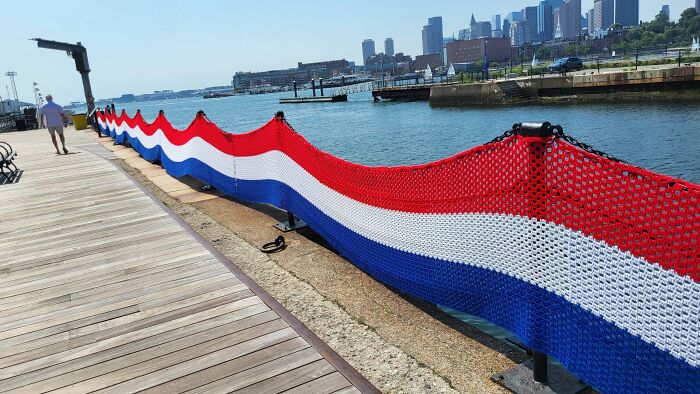 A long, festive red, white, and blue crochet banner adorns a waterfront railing with a city skyline in the background, an incredible crochet display.