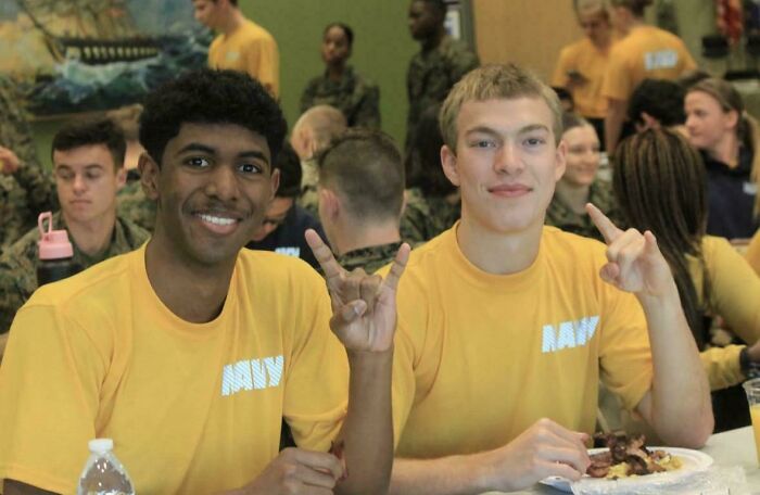 Two young men, one with dark curly hair and one with blond hair, posing with hand gestures in a real life optical illusion.