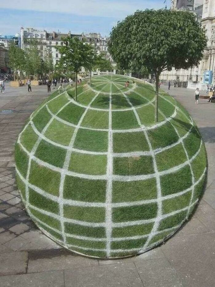 A large, spherical optical illusion of grass with white grid lines, resembling a globe with trees on top, in a city square.