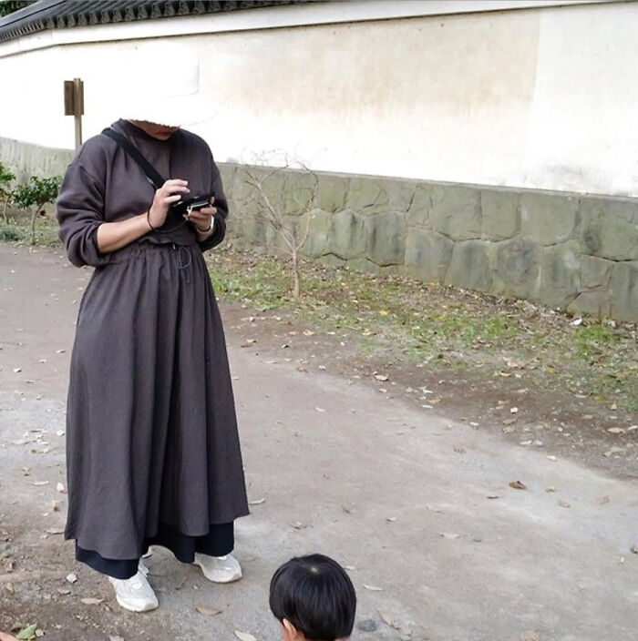 A woman wearing a long dark dress and white shoes, looking at her phone, creating an optical illusion with a small child.