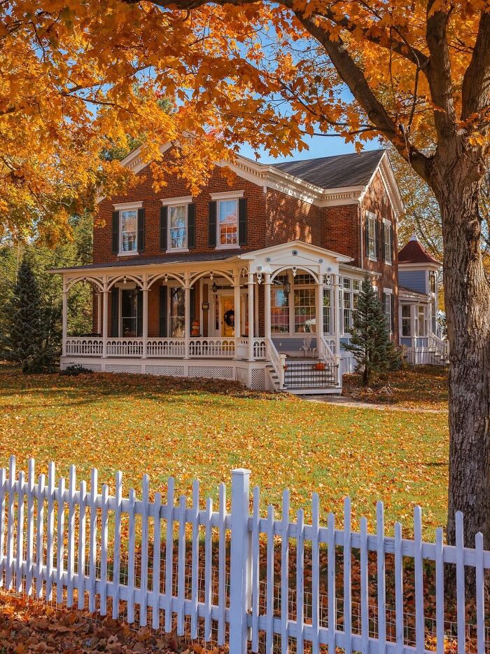 A classic brick house with beautiful architecture, a white picket fence, and vibrant autumn leaves scattered across the lawn.