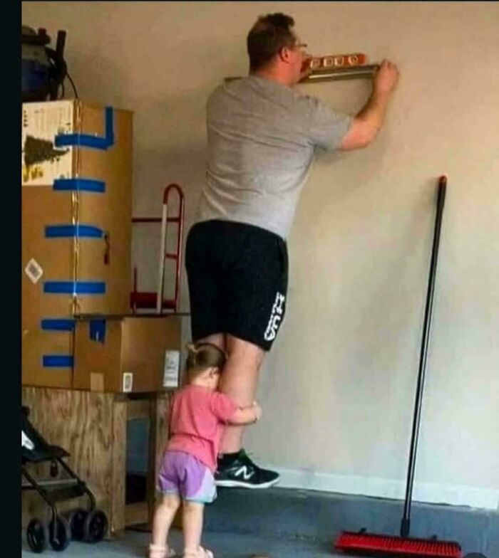 A little girl hugs her dad's leg as he works on the wall, creating an optical illusion of scale.