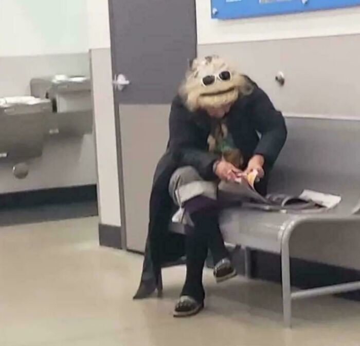 A person wearing a furry, monster-like headpiece while sitting on a bench, an optical illusion.