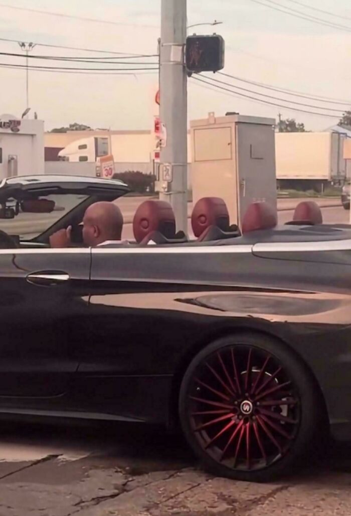 A black convertible car with passengers whose bald heads align with the red headrests, an optical illusion.