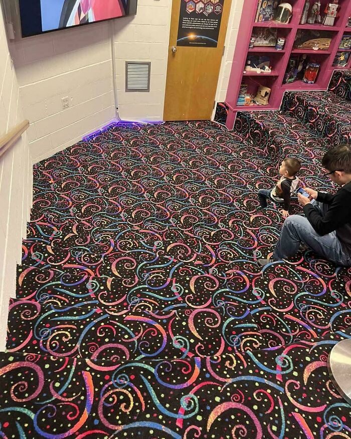 A man and child sit on a brightly patterned carpet that creates an optical illusion of uneven surfaces, a real-life optical illusion.
