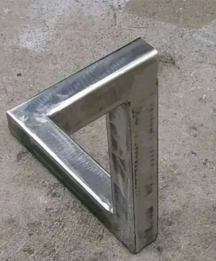 A metal optical illusion triangle, appearing to be impossible, rests on a concrete surface, showcasing a real-life optical illusion.