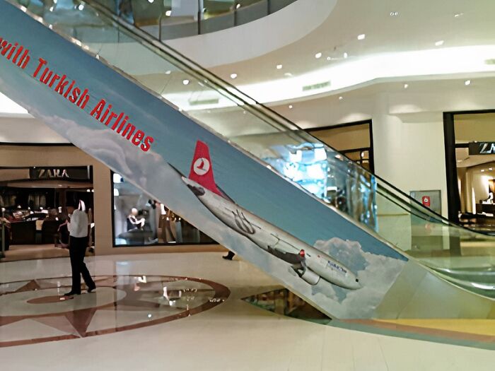 An escalator in a mall with a Turkish Airlines advertisement featuring an airplane, but the design makes it look like the plane is crashing due to a designer's failed job.