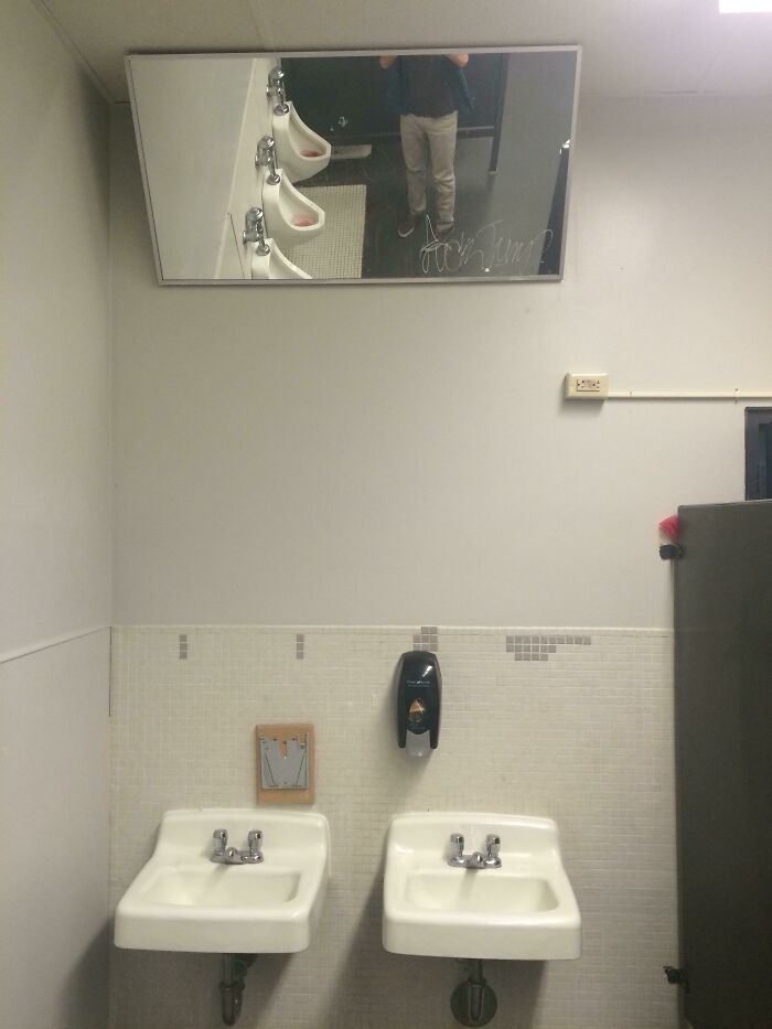 A public restroom with a mirror reflecting urinals and a person's legs, an example of designers failing dismally.