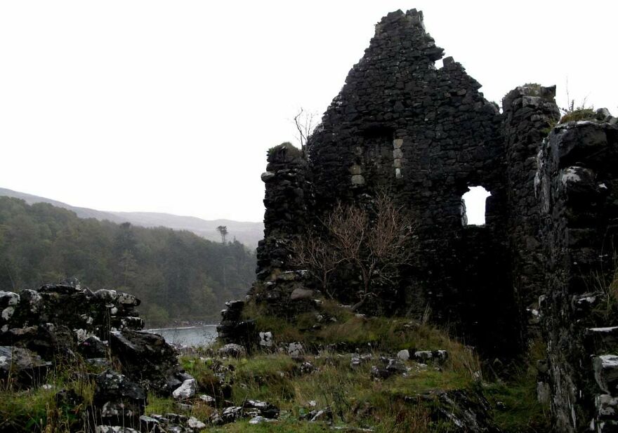 Ruins of an old, interesting castle made of dark stone, overlooking a misty body of water and forested hills.