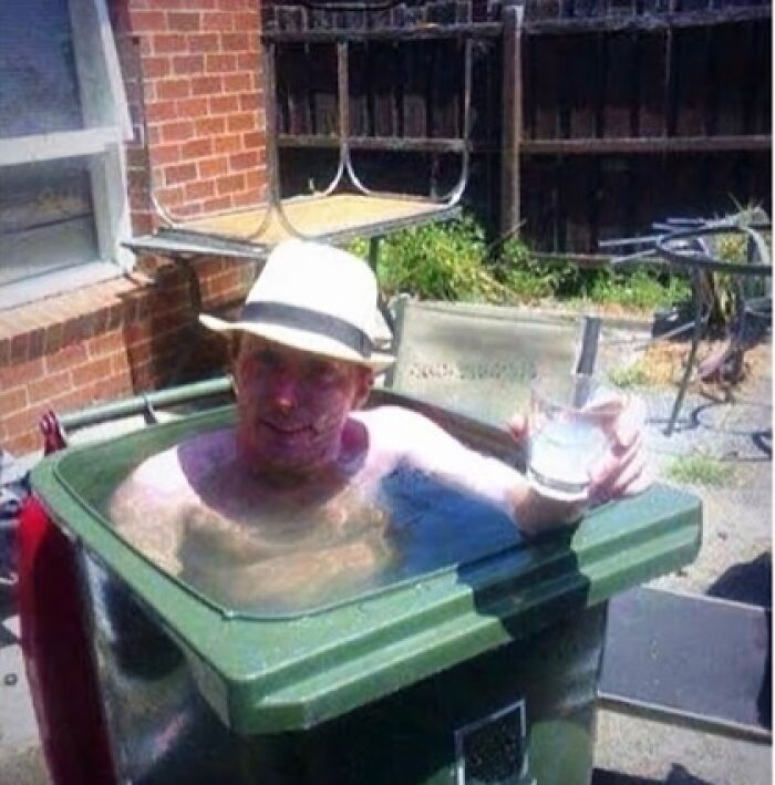Funny meme about summer heat with a man in a fedora sitting in a trash can filled with water, holding a drink.