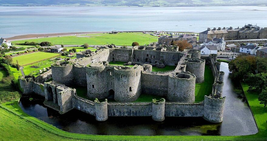 An aerial view of a large, ancient stone castle with a moat, surrounded by green fields and a distant body of water, creating a fairytale scene.