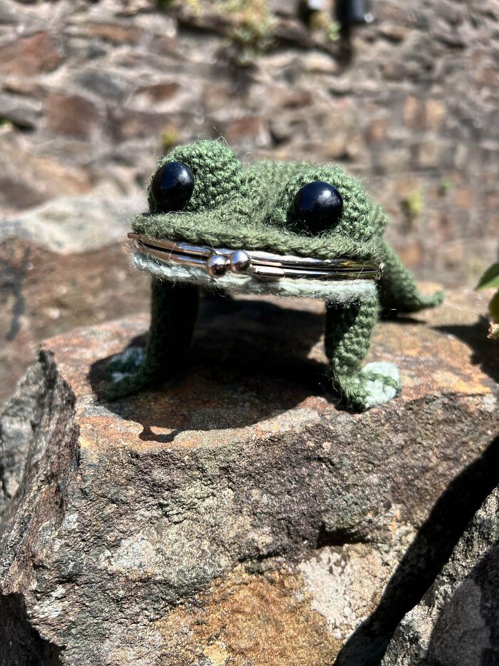 A charming green crochet frog coin purse with large black eyes sits on a rock, an incredible crochet creation.