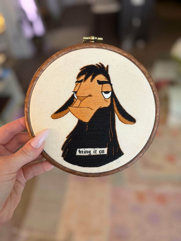 A hand holding a circular embroidery hoop with a stitched cartoon llama design and the text bring it on