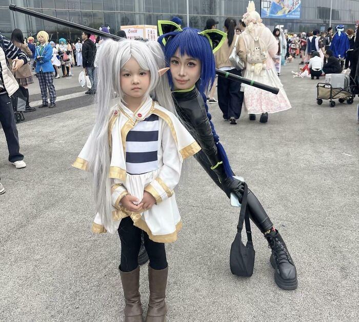 A child in a white wig standing in front of a person in a blue wig, creating an optical illusion.
