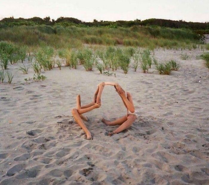 Two sets of human limbs on a sandy beach create an optical illusion appearing as a single, abstract figure.