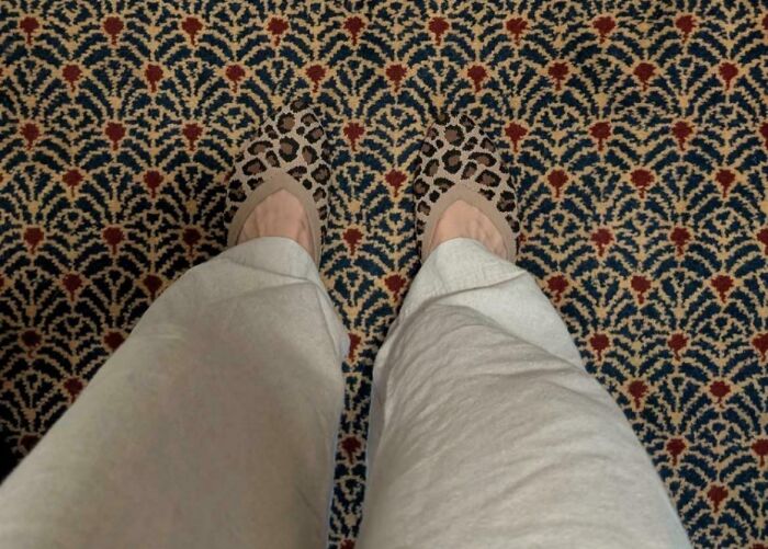A pair of feet with leopard print shoes blending seamlessly into an optical illusion on patterned carpet.