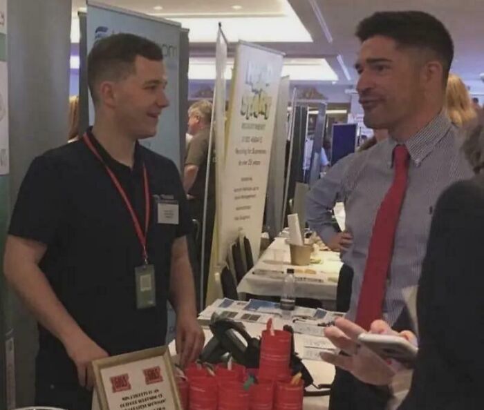Two men in business attire at a networking event, creating an optical illusion with a perspective trick.