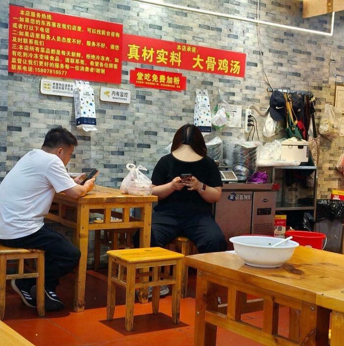 Two people at a table, both engrossed in their phones, creating a subtle optical illusion in the restaurant setting.