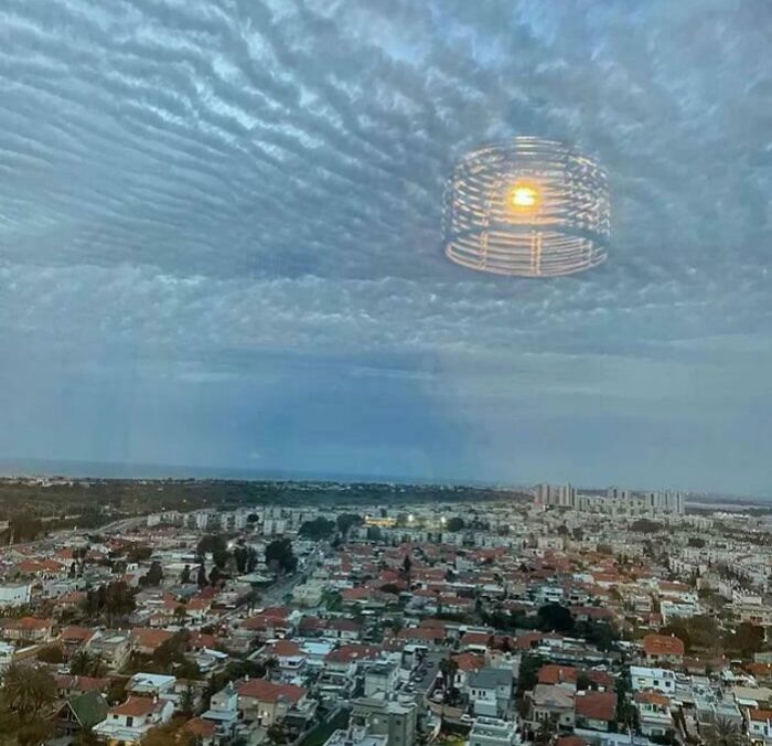 An optical illusion of a lamp appearing as a giant UFO in the sky over a city.