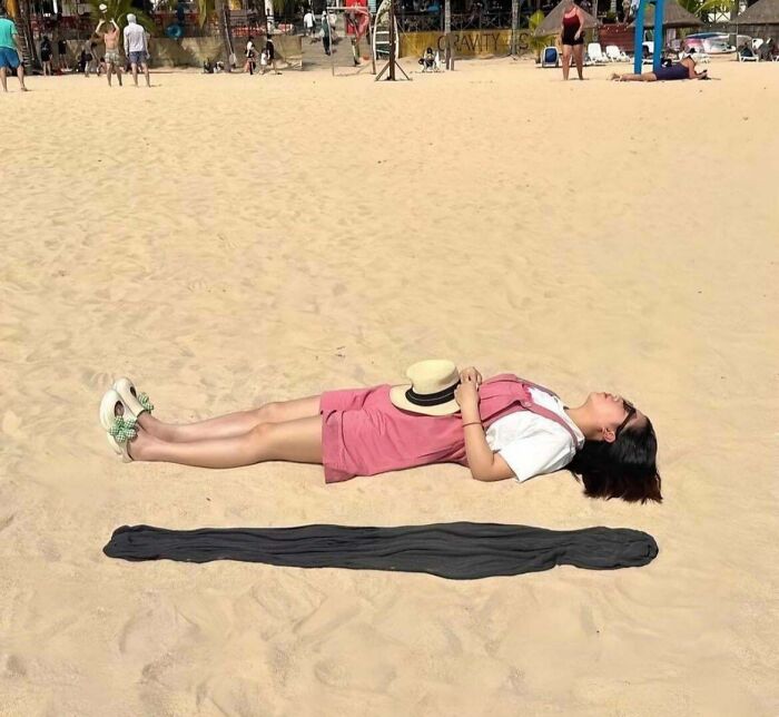 A woman lying on a sandy beach with a hat on her stomach, casting a long, dark shadow that forms an optical illusion.