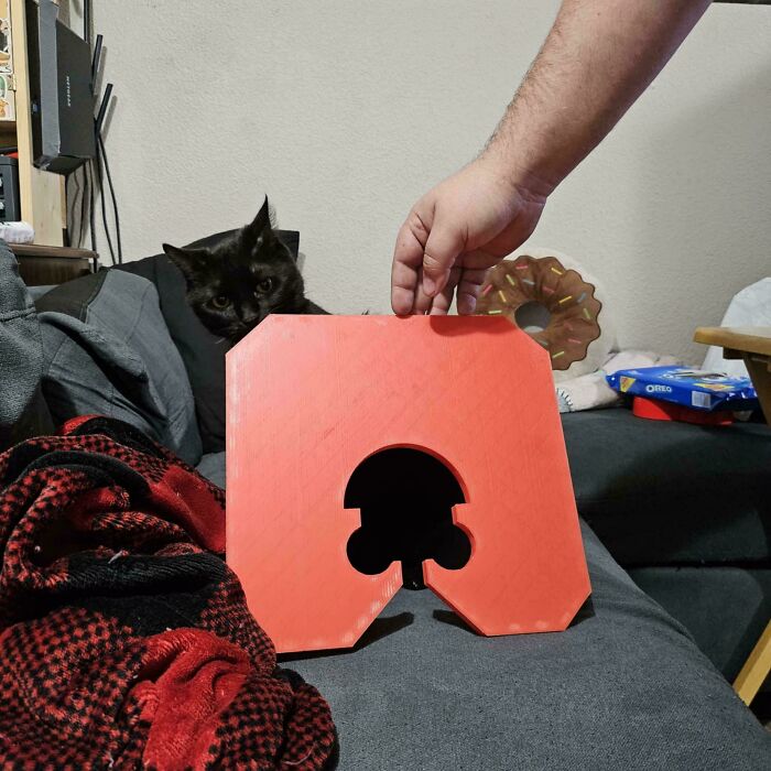 A hand presenting a red, custom-designed 3D printer object to a curious black cat, a cool 3D printer project.