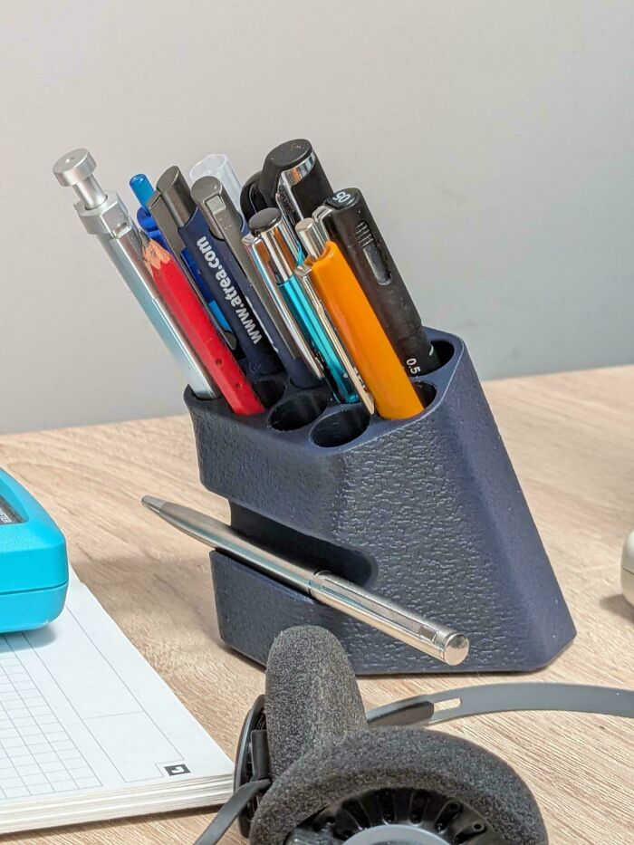 A functional 3D printer project, a dark blue textured pen holder filled with various pens and pencils on a wooden desk.
