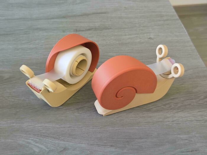 Two charming 3D printer projects of snail-shaped tape dispensers, one with tape loaded and the other empty, on a wooden surface.
