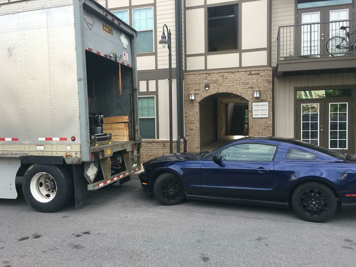 A blue Ford Mustang parked behind a moving truck, blocking the ramp. An entitled man ignores boundaries.