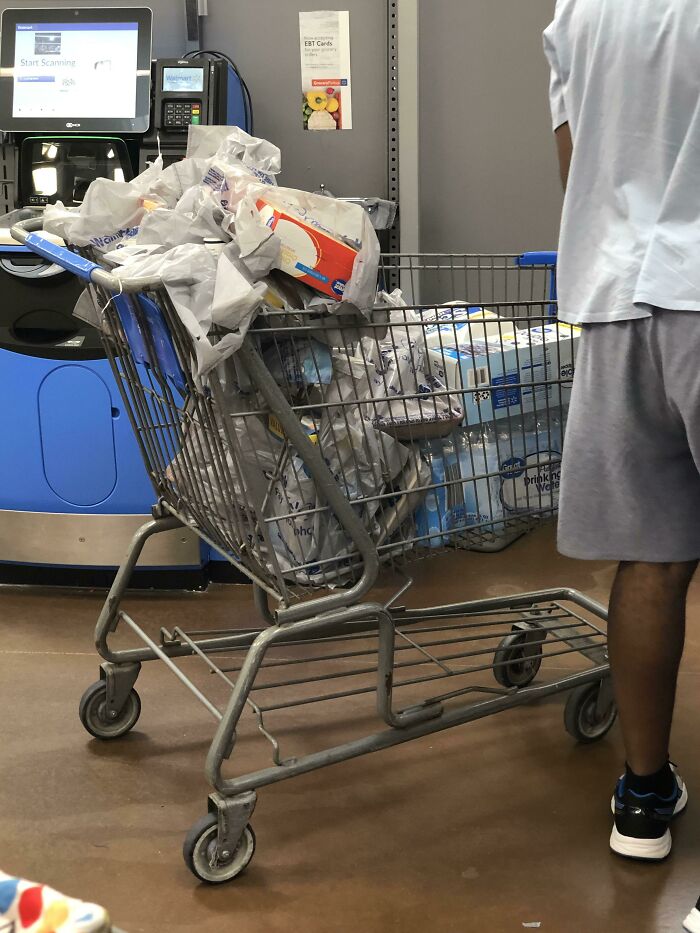 A shopping cart overflowing with groceries, blocking a self-checkout aisle. An entitled man shows no boundary awareness.