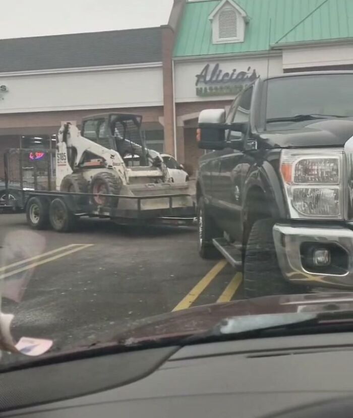 A black truck towing a bobcat, parked carelessly near a store, illustrating entitled men.