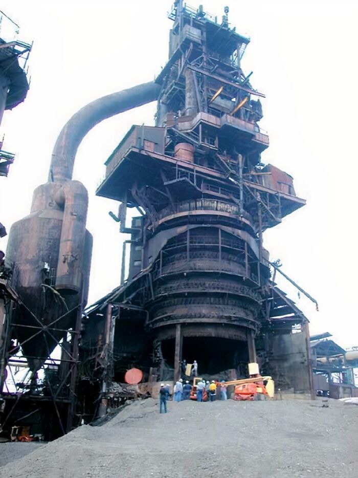 Workers dwarfed by an extremely large, decaying industrial blast furnace, highlighting the scary and eerie scale.