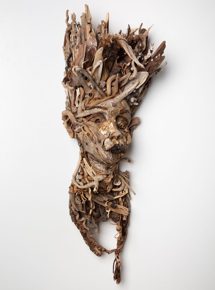 Elongated wood sculpture of a face and upper torso made from collected natural wood pieces, against a white background.