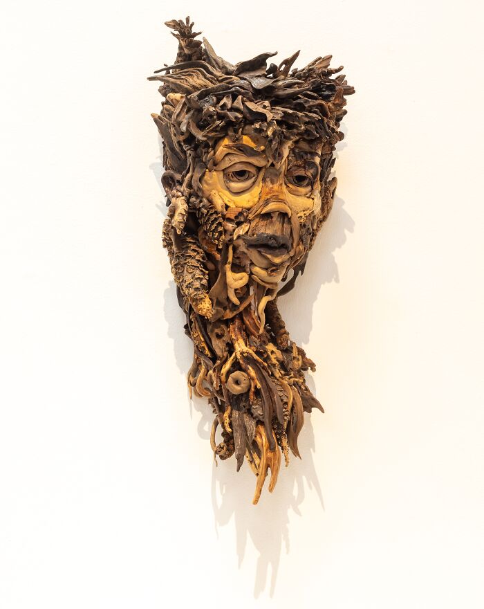 A detailed wood sculpture of a somber face and neck, created from collected pieces of wood, mounted on a white wall.