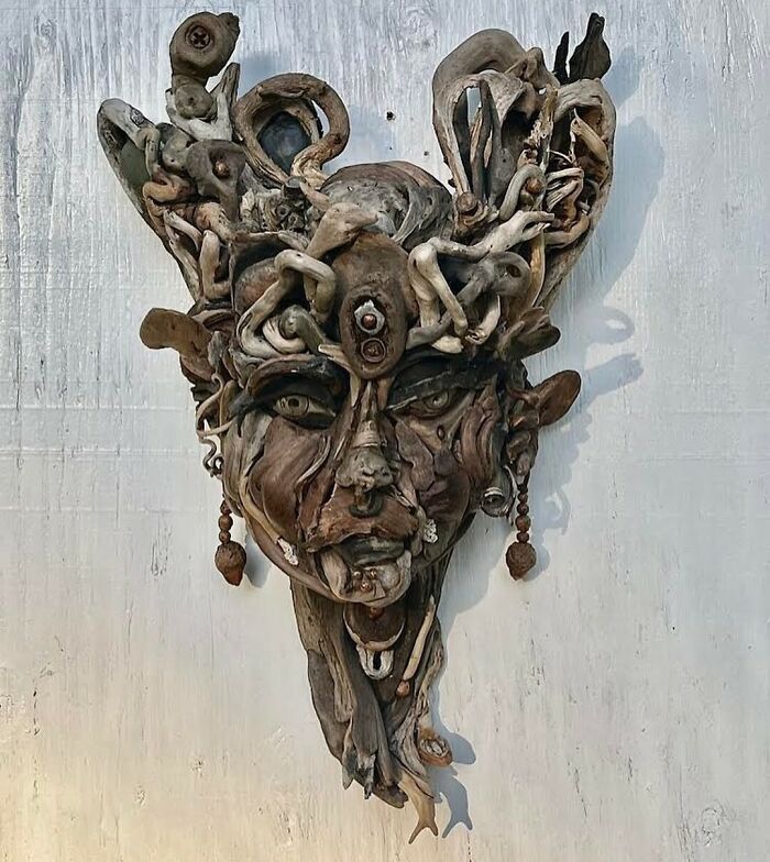Detailed wood sculpture of a face with elaborate hair made from collected natural pieces, hanging on a white wall.