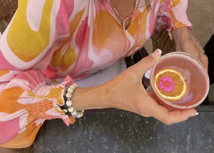 A person holding a drink with an orange slice and pink flower, wearing a shirt that perfectly matches the drink and flower, a satisfying paint match.